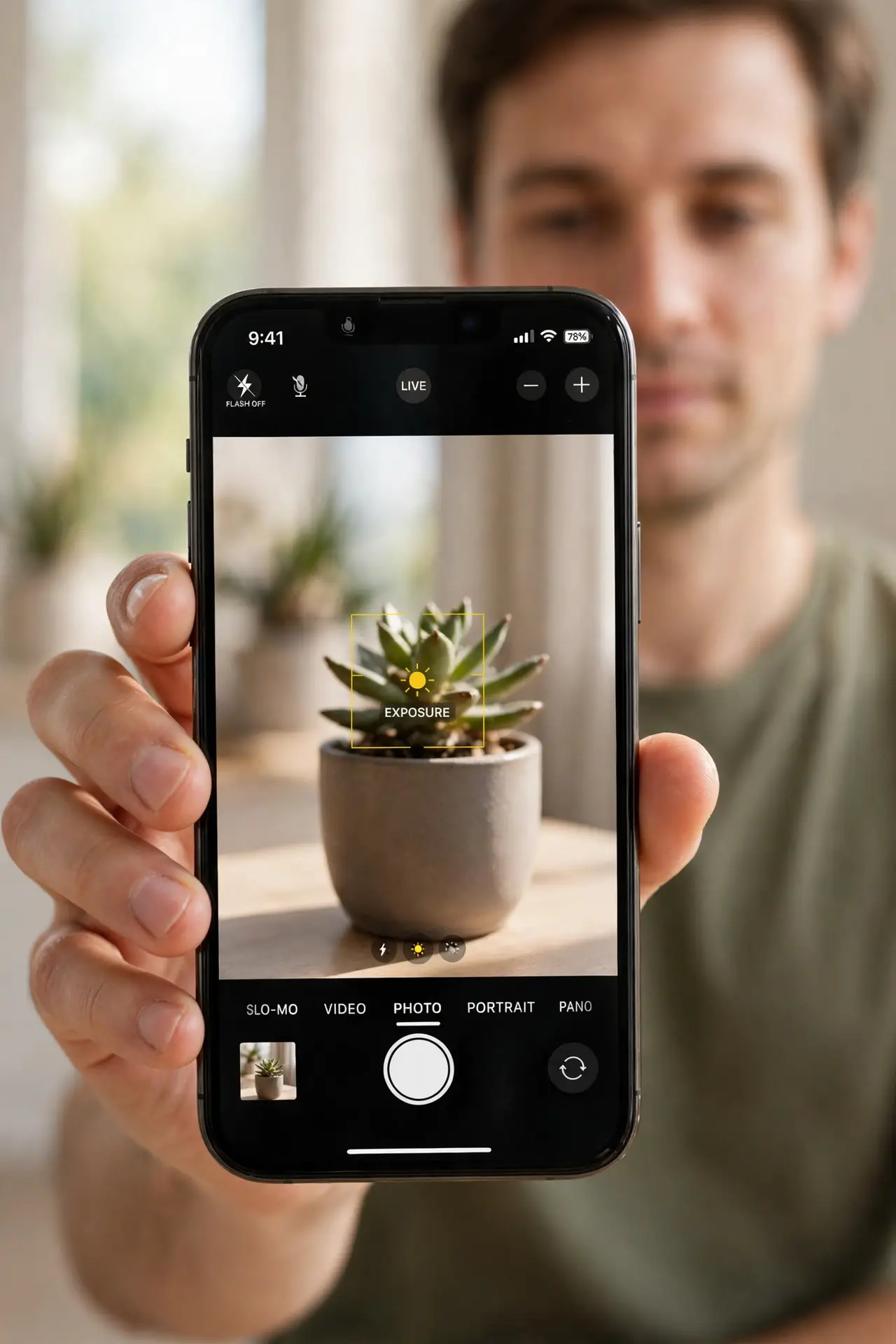 Photorealistic photo: A calm adult holding an iPhone toward the viewer, the real iPhone screen showing the Camera app with a subject in sharp focus and a focus box legible.